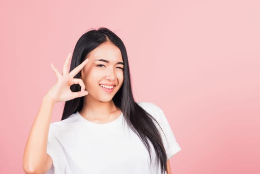 Portrait Asian beautiful young woman made finger OK symbol sign gesture wink eyes to agree and looking at camera, studio shot isolated on pink background, Thai female agree hand sign copy space