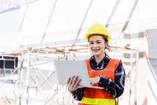Asian engineer architect worker woman holding laptop inspect and oversee infrastructure progress at construction site, engineering use computer to operate project and control worker to building