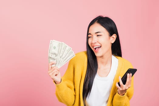 Portrait Asian happy beautiful young woman teen shopper smiling standing excited holding online smart mobile phone and dollar money banknotes on hand in summer, studio shot isolated on pink background