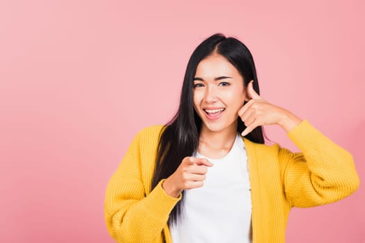 Portrait Asian beautiful young woman smiling doing phone gesture with hand fingers on telephone and pointing to camera while making call back to me gesture, studio shot isolated pink background