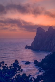 Beautiful seascape at sunset with clouds in the sky. In the foreground are dark branches of trees, the sea reflects the lilac sunset sky, the mountain is also in a sunset haze of heavenly color.