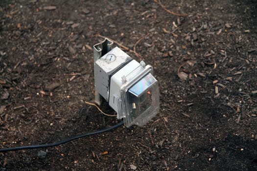 Outdoors public electric outlet with rain cover and a black plugged cable emerging from the brown soil