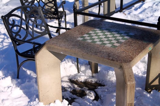 A concrete chessboard table with a heavy iron chair in the snow in the park