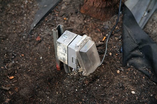 Outdoors public electric outlet with rain cover and a black plugged cable emerging from the brown soil