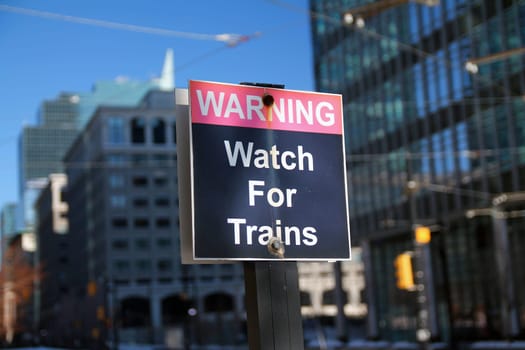 Warning Watch for Trains black and red sign with buildings on the back and reflecting the sun
