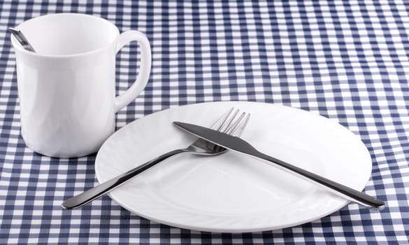 The Empy Mug with spoon, plate with a knife and fork on the tablecloth in a cage. View from above with copy space