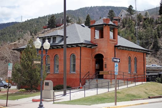 Idaho Springs, CO, USA - April 19, 2014: Front and side view of a brick city hall building with bell tower at 1711 Miner Street in downtown. Brick City Hall building with ramp walkway, parking, Highway 70, homes and evergreen tree covered mountains in the background on a warm spring day.