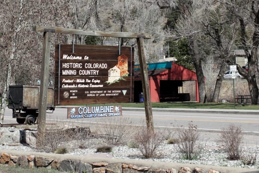 Idaho Springs, CO, USA - April 21, 2014: Old wooden sign welcoming visitors to historic Colorado mining country with other information. The sign has a painting of the towns Argo Mine as well as an antique mining cart behind the sign on a sunny spring day.