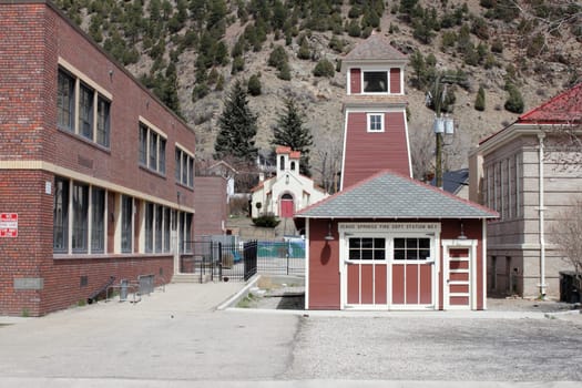 Idaho Springs, CO, USA - April 19, 2014: Historical architecture rises up with a bell on top of this old and small, reddish brown fire station located in the west part of downtown. A rare antique fire station with old fashioned doors next to a school and the main library, near a picturesque church all with various shades of red in front of a sunny mountain background.
