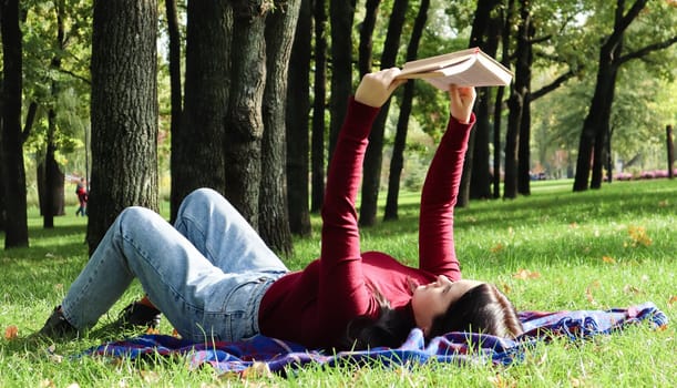 Young woman lying and reading her favorite book on a woolen plaid blanket in a city park on green grass on a pleasant sunny day. The concept of recreation, education and remote study