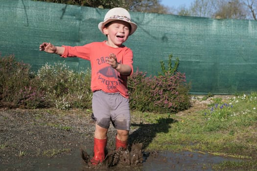Happy male kid in rubber boots have fun playing in a puddle mud