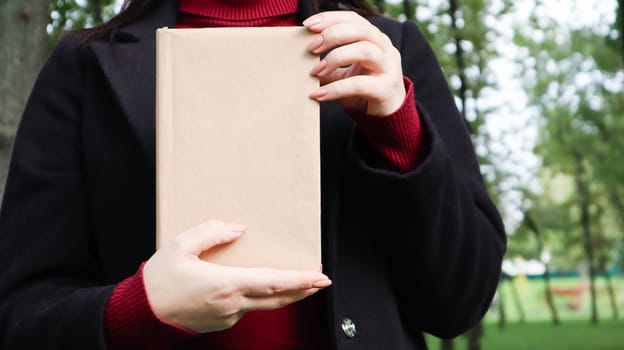Close up view of woman in black woolen coat and burgundy sweater holding a book with empty cover in the park. Free space for your book reading concept background layout