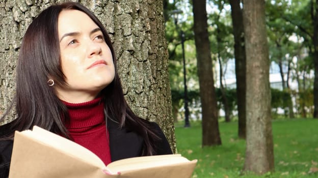 Portrait of a beautiful young woman who is sitting under a tree and reading her favorite book in a city park on green grass on a pleasant sunny day. Vacation, education and study concept