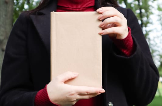 Close up view of woman in black woolen coat and burgundy sweater holding a book with empty cover in the park. Free space for your book reading concept background layout