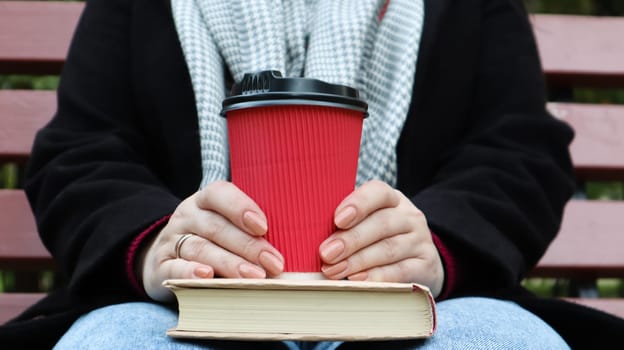 Young woman in jeans, coat and scarf, on a park bench. A woman is reading a book and drinking coffee or other hot drink outdoors alone. Close-up. The concept of honor, study, leisure and recreation
