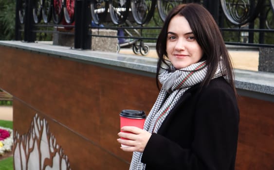 Young stylish woman in a coat and scarf drinks morning hot coffee in a red eco paper glass outdoors in an autumn park. Portrait of a young woman holding a takeaway coffee cup, shallow depth of field.