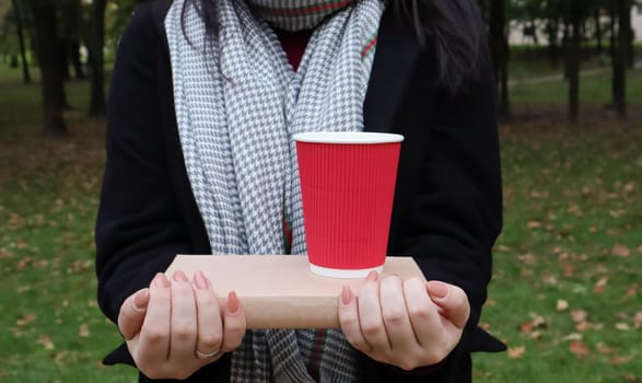 A young woman in a black coat and a gray scarf holds one book and a glass with coffee in her hands on the background of an autumn park. On open air. Education, close hands of the girl