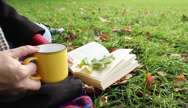 A girl sits on a blanket in the autumn forest, reads a book and drinks a hot drink from a yellow cup in a city park on a warm day on a green meadow. The concept of reading, relaxation and comfort