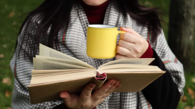 A girl dressed in a coat and a scarf in the autumn forest holds a book and a cup with a hot drink in her hands close-up in a city park on a warm day. The concept of reading, relaxation and comfort