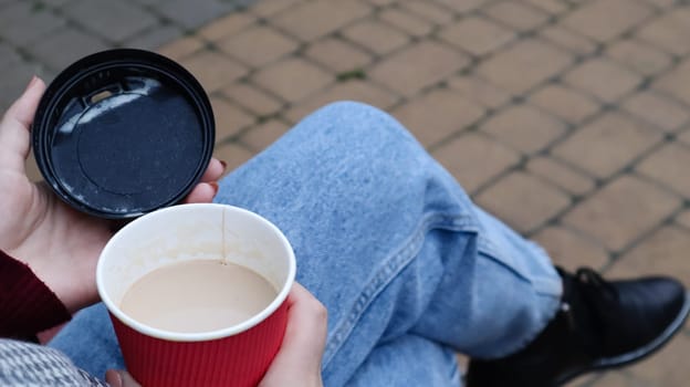 Female hands are holding morning takeaway coffee and closing a paper cup with a lid while sitting on a park bench, close-up of hands