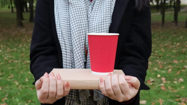 A young woman in a black coat and a gray scarf holds one book and a glass with coffee in her hands on the background of an autumn park. On open air. Education, close hands of the girl