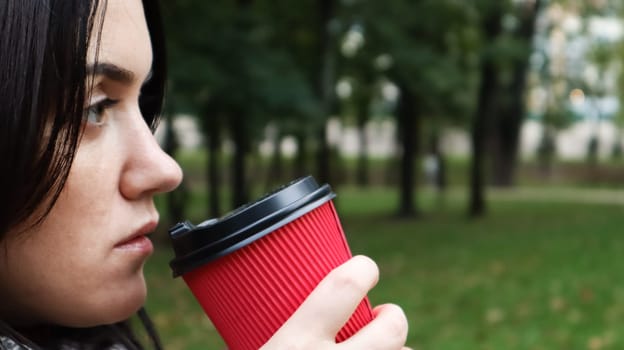 Young stylish woman drinks morning hot coffee in red eco paper cup outdoors in autumn park. Close-up side view of young woman holding takeaway coffee cup