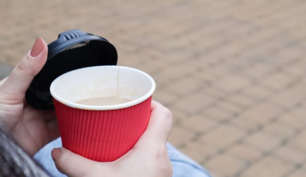 Female hands are holding morning takeaway coffee and closing a paper cup with a lid while sitting on a park bench, close-up of hands
