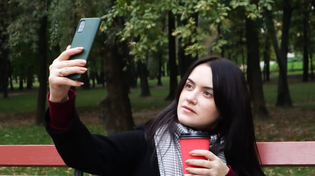 Beautiful young caucasian woman in a coat with a paper cup of takeaway coffee taking a selfie or taking pictures of herself for a blog with a smartphone outdoors in a park sitting on a bench