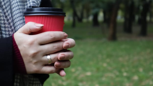 A young girl in a coat holds morning coffee with her while walking in the park. Hand holding paper cup of coffee in green park. Takeaway cappuccino. Close-up, copy space