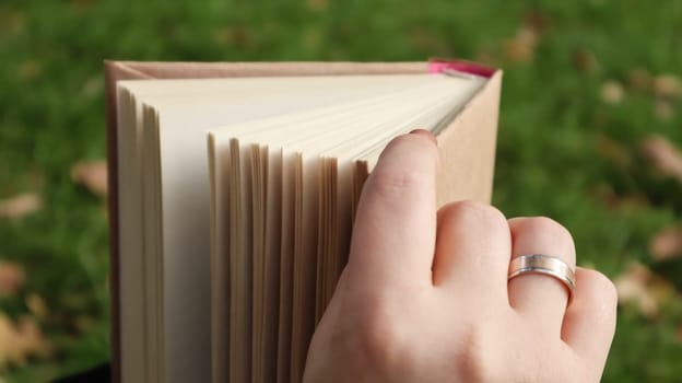 Girl reading a book in the autumn park. Female hands open the pages of a paper book outdoors on a warm sunny day. The student is preparing for the exam. Literary leisure in nature