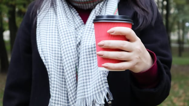 A young girl in a coat holds morning coffee with her while walking in the park. Hand holding paper cup of coffee in green park. Takeaway cappuccino. Close-up