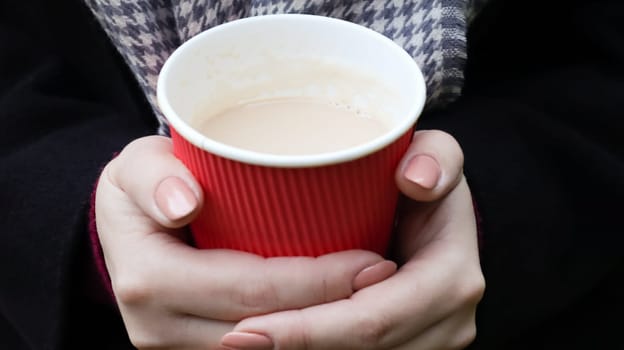 A young girl in a coat holds morning coffee with her while walking in the park. Hand holding paper cup of coffee in green park. Takeaway cappuccino. Close-up