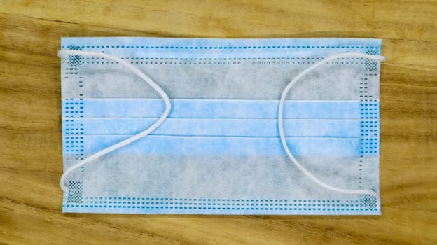 One 3-ply disposable surgical face mask with rubber pads covering the mouth and nose on a wooden table. The concept of protection from bacteria, healthcare and medicine. COVID-19. Dangerous virus
