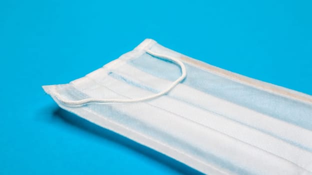 One 3-ply disposable surgical face mask with rubber grips covering mouth and nose against blue background. Protection from bacteria, healthcare and medicine concept. COVID-19. A dangerous virus