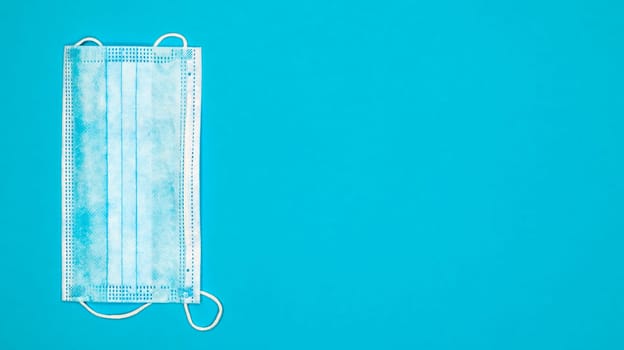 One 3-ply disposable surgical face mask with rubber pads covering the mouth and nose against a blue background. The concept of protection from bacteria, healthcare and medicine. COVID-19. copy space.