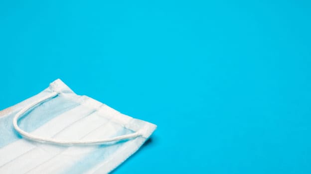 One 3-ply disposable surgical face mask with rubber pads to cover mouth and nose against blue background. The concept of protection from bacteria, healthcare and medicine. COVID-19. copy space