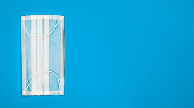 One 3-ply disposable surgical face mask with rubber pads to cover mouth and nose against blue background. The concept of protection from bacteria, healthcare and medicine. COVID-19. copy space
