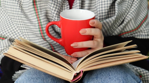 A girl dressed in a coat and a scarf in the autumn forest holds a book and a cup with a hot drink in her hands close-up in a city park on a warm day. The concept of reading, relaxation and comfort