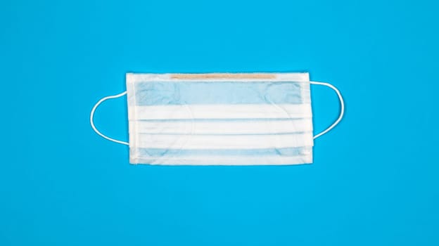 One 3-ply disposable surgical face mask with rubber grips covering mouth and nose against blue background. Protection from bacteria, healthcare and medicine concept. COVID-19. A dangerous virus