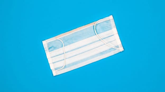 One 3-ply disposable surgical face mask with rubber grips covering mouth and nose against blue background. Protection from bacteria, healthcare and medicine concept. COVID-19. A dangerous virus