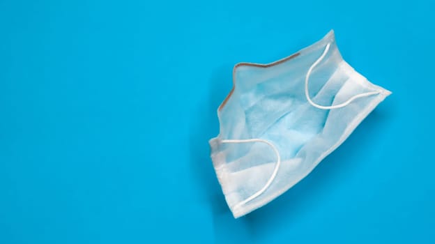 One 3-ply disposable surgical face mask with rubber pads covering the mouth and nose against a blue background. The concept of protection from bacteria, healthcare and medicine. COVID-19. copy space.