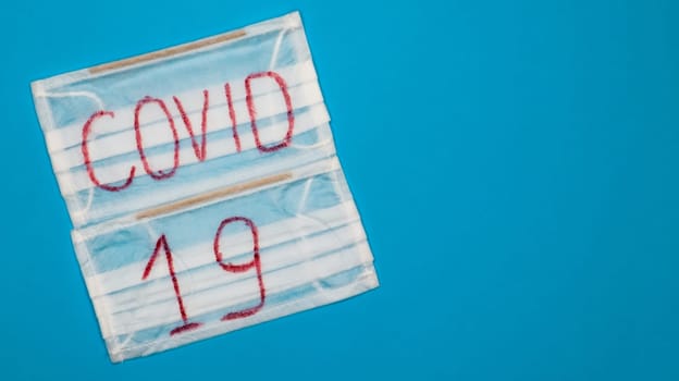 Typical white 3-ply surgical mask with rubber earhooks to cover mouth and nose with English block letters Covid-19 on a blue background. Dangerous virus and quarantine concept. Copy space.