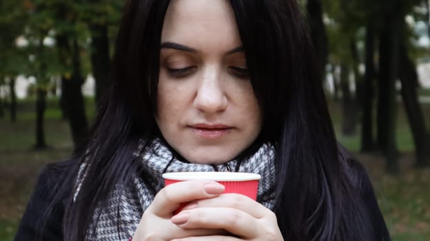 Portrait of a young beautiful woman in a coat who holds morning coffee with herself near her lips while walking in the park. Hand holding paper cup of coffee in green park. Close-up