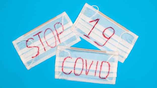 Typical 3-ply white surgical mask with rubber earhooks to cover mouth and nose with English block letters Stop Covid-19 on a blue background. Dangerous virus and quarantine concept.