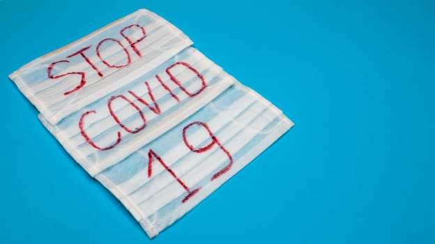 Typical 3-ply white surgical mask with rubber earhooks to cover mouth and nose with English block letters Stop Covid-19 on a blue background. Dangerous virus and quarantine concept. Copy space.