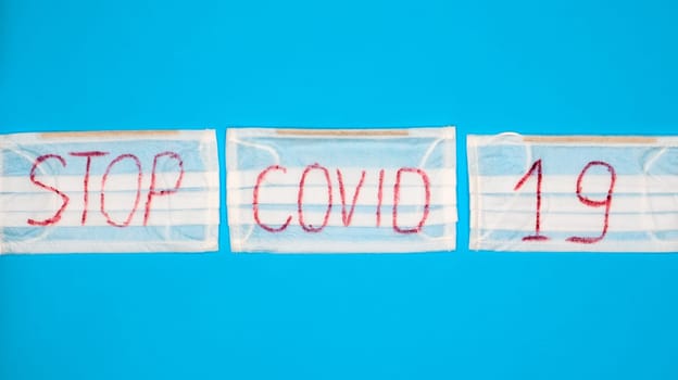 Typical 3-ply white surgical mask with rubber earhooks to cover mouth and nose with English block letters Stop Covid-19 on a blue background. Dangerous virus and quarantine concept. Copy space.