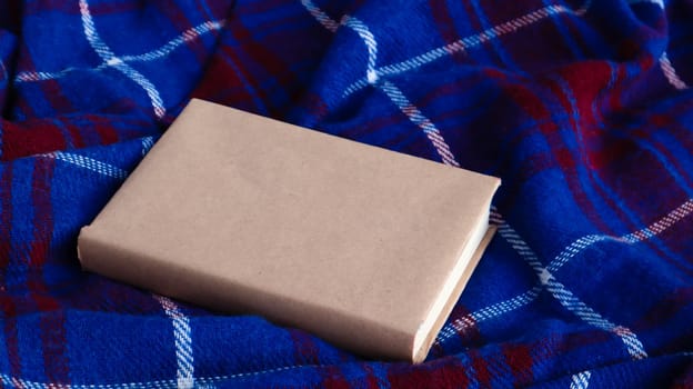 Cozy still life. A closed book with a warm blue and red woolen plaid blanket. Free copy space. Winter and autumn layout with blank book cover. Warm festive background