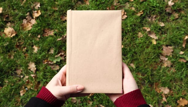 A closed book in a cover made of craft paper in female hands with green grass and fallen yellow leaves in the background. Flat lay, top view. Template, layout. Copy space