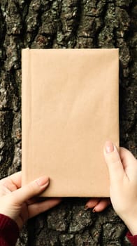 A closed book in a cover made of craft paper in female hands on the background of a tree bark in the park. Copy space. The concept of reading, recreation and leisure, study and education