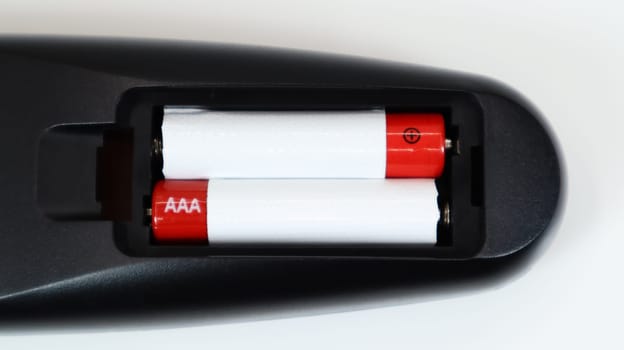 Black TV remote control with AAA alkaline batteries in red and white on a white background. Battery replacement, spare parts. Remote control battery compartment close-up
